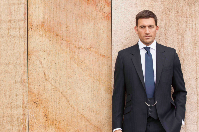 A well-dressed man in a suit and tie leans against a wall, showcasing a relaxed yet polished demeanor