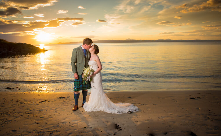 A bride and groom share a kiss on the beach during a vibrant sunset, creating a romantic and serene atmosphere.